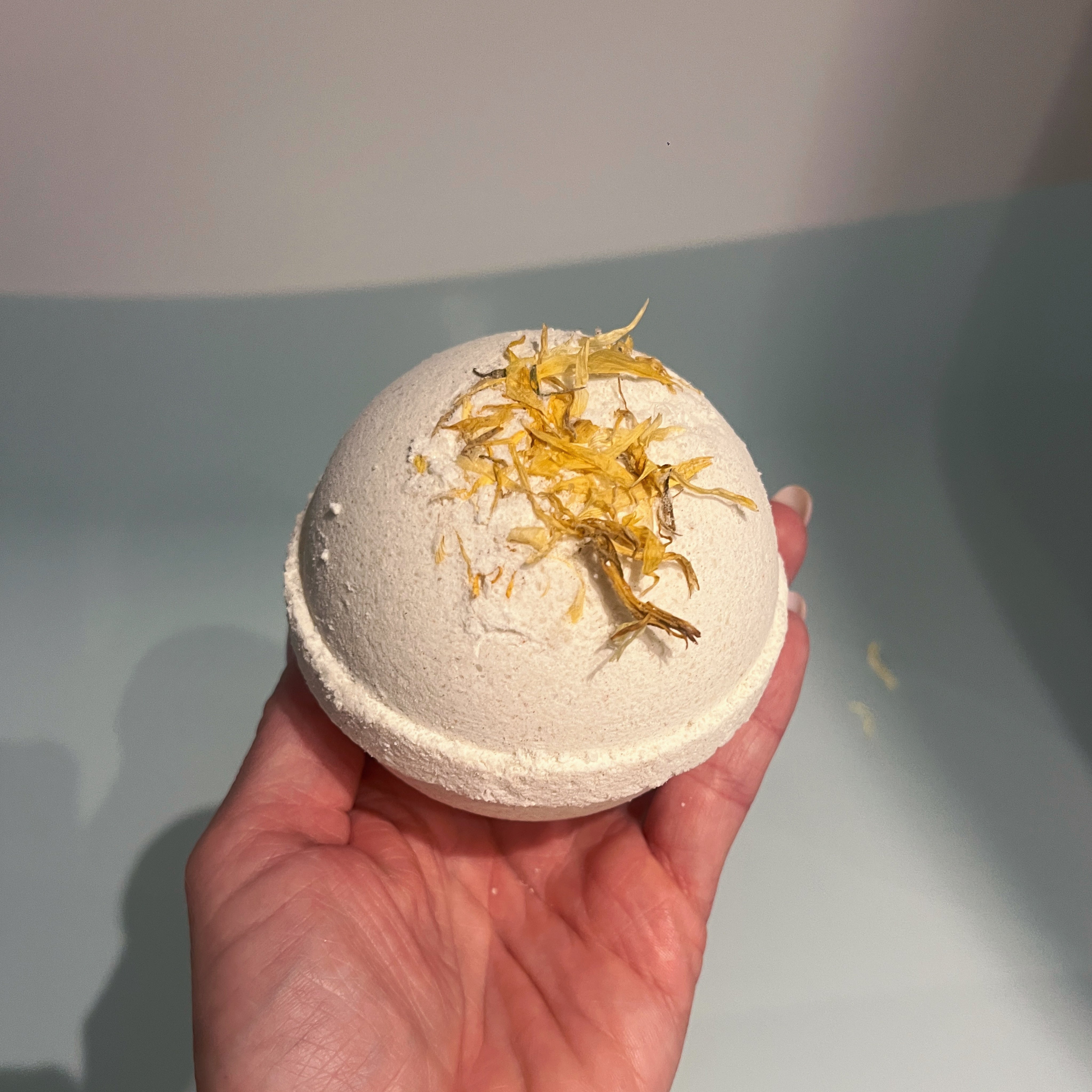 Energy Balance bath bomb with vanilla, citrus and calendula leafs slowly placed into the warm water of a white bathtub.